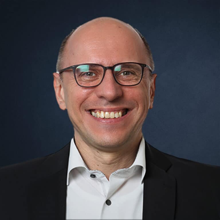 Porträt eines Mannes mit Brille und lächelndem Gesicht, in einem schwarzen Anzug und weißem Hemd.