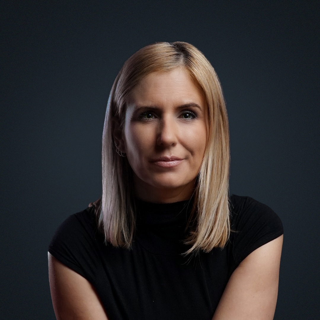 Porträt einer Frau mit blonden Haaren und schwarzem Oberteil vor dunklem Hintergrund.