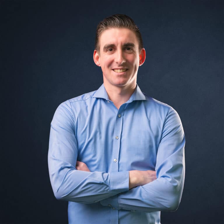 Porträt eines Mannes mit blauer Hemd, der mit verschränkten Armen lächelt.