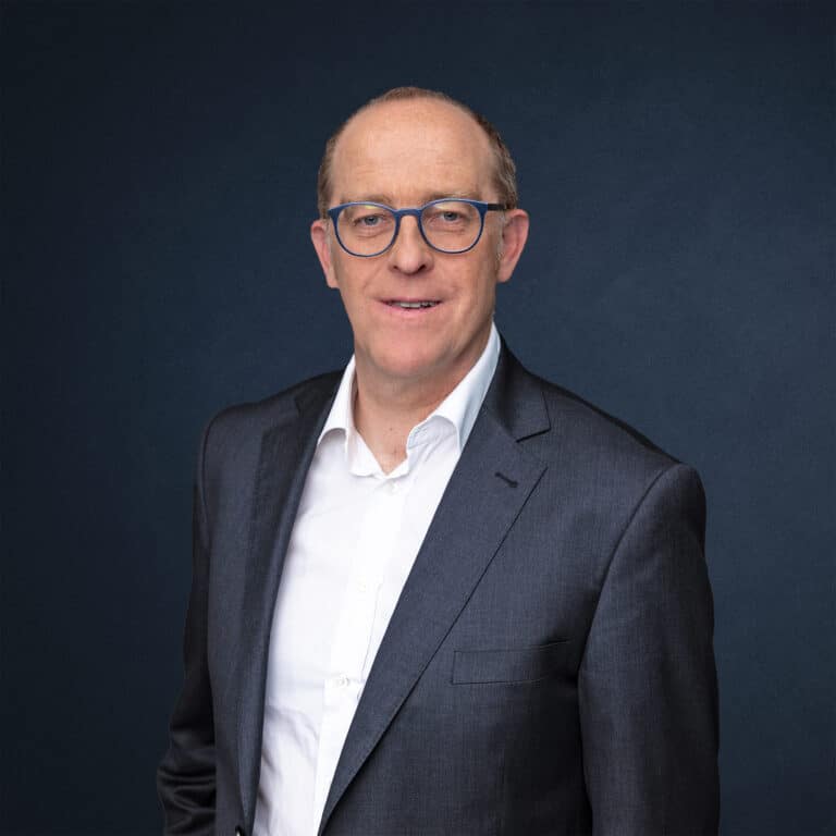 Michael Becker trägt einen Anzug und eine Brille, steht vor einem dunklen Hintergrund.