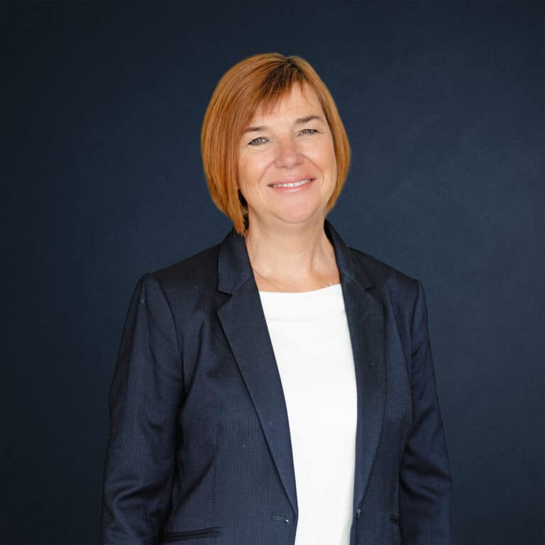 Karin Freischem mit orangefarbenem Haar, trägt einen dunkelblauen Anzug und lächelt vor einem dunklen Hintergrund.