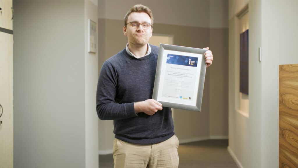 Ein Mann hält ein gerahmtes Zertifikat in der Hand und steht in einem Innenraum.