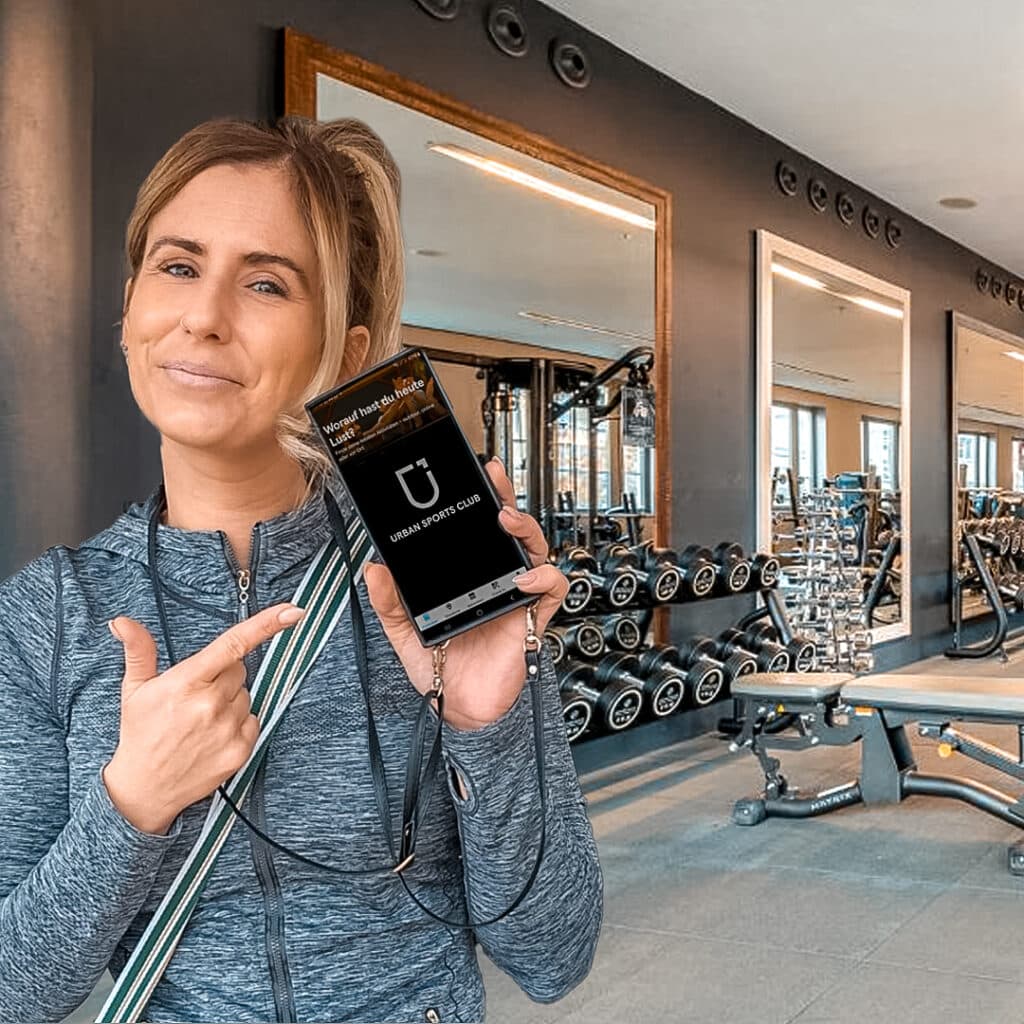 Frau zeigt auf ihr Smartphone im Fitnessstudio mit Gewichten im Hintergrund.