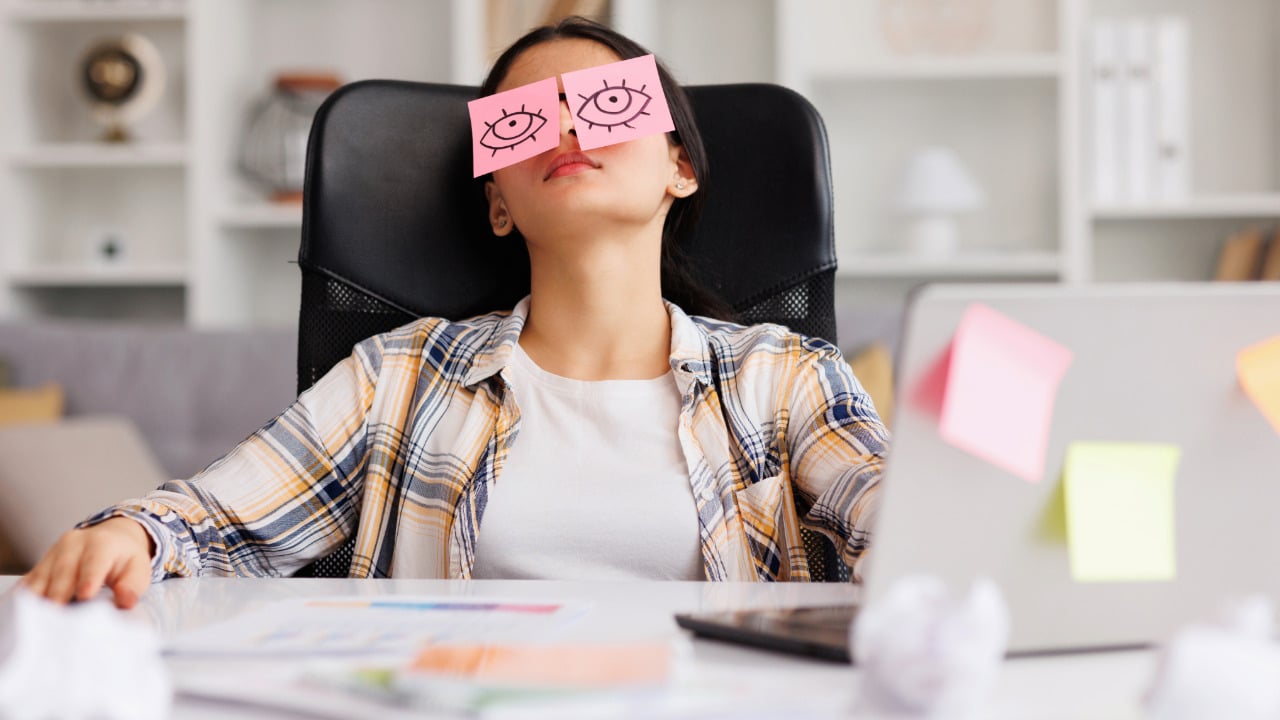 Frau sitzt an einem Schreibtisch mit Post-it-Zetteln auf den Augen, umgeben von Notizen und einem Laptop.
